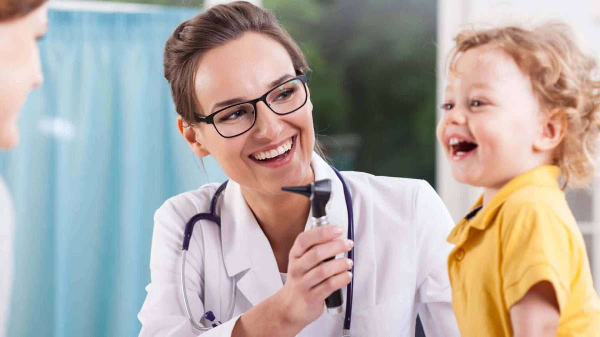 Ärztin mit Brille hält ein Otoskop und lächelt ein lachendes Kind an.